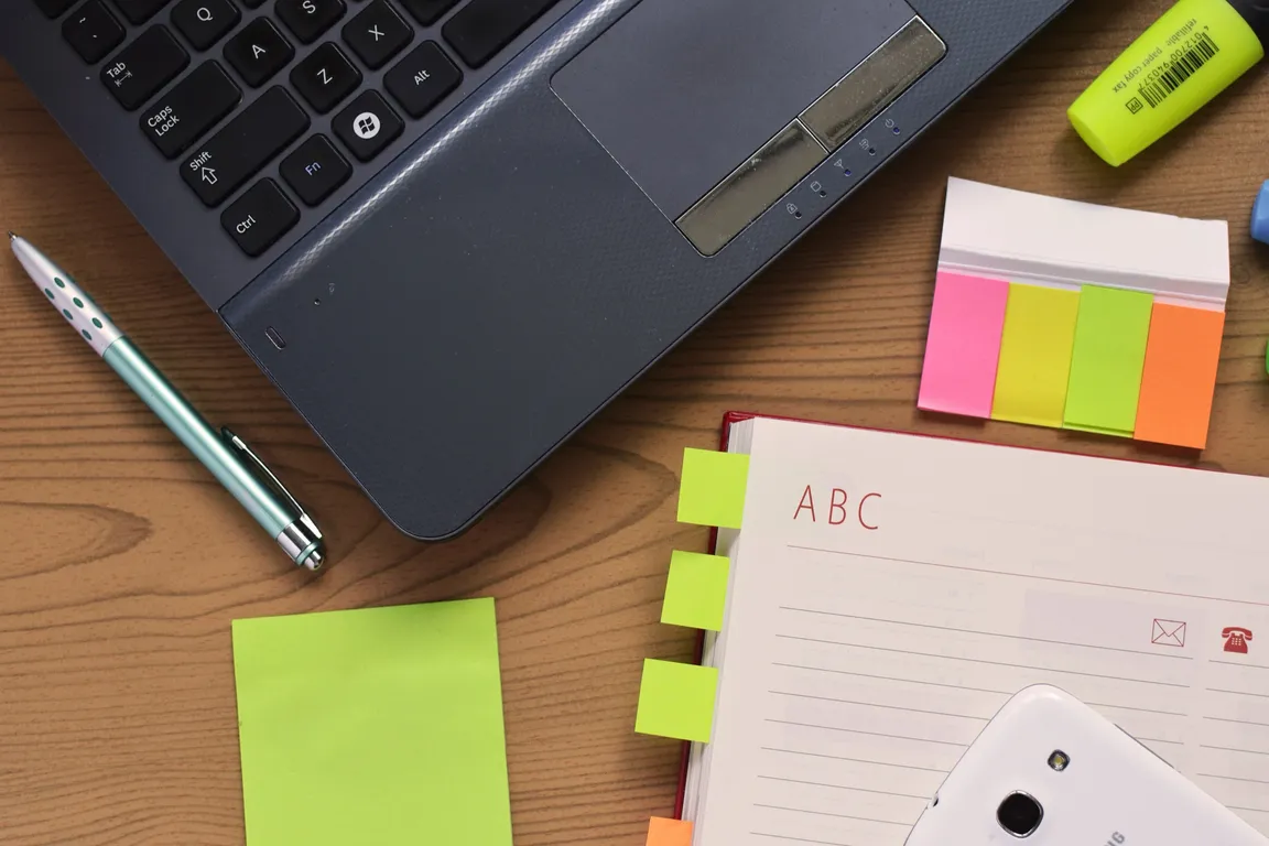 An open address book with alphabet tabs lying on a wooden desk beside a laptop, a pen, sticky notes and a highlighter.
