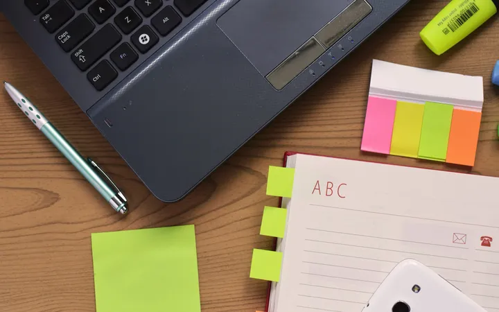 An open address book with alphabet tabs lying on a wooden desk beside a laptop, a pen, sticky notes and a highlighter.