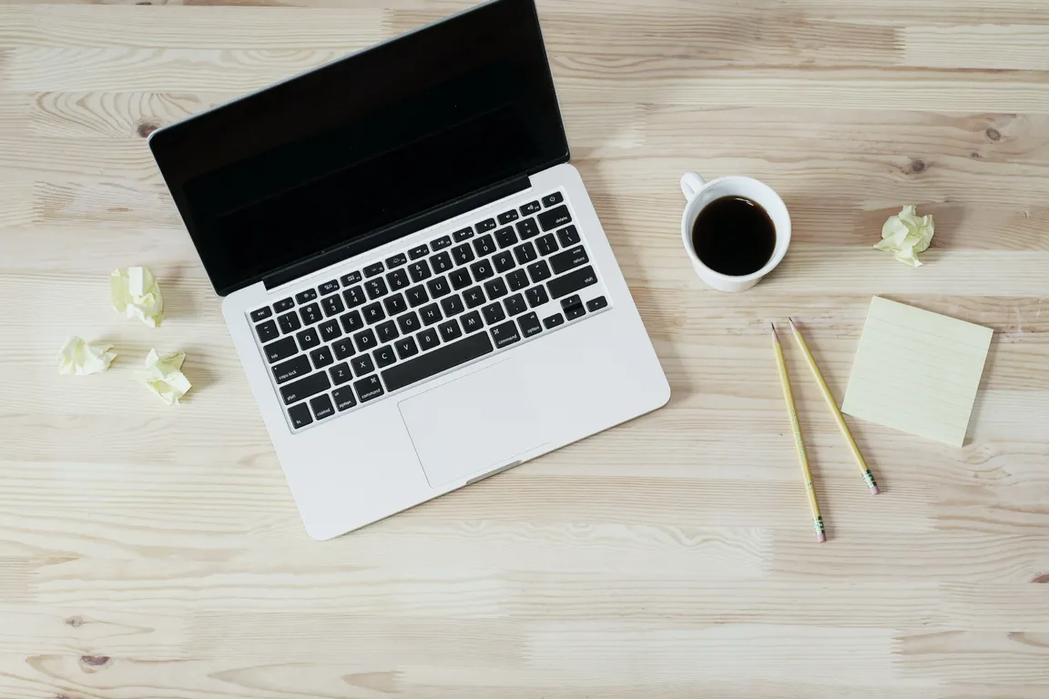 A laptop seen from above on a pale wooden desk, next to a cup of black coffee, two pencils, a pad of sticky notes and a few crumpled paper notes.